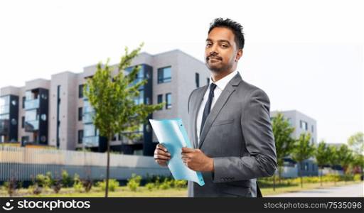 real estate business and people concept - indian man realtor with folder over living houses on city street background. indian man realtor with folder on city street
