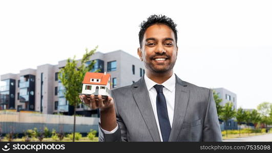 real estate business and people concept - indian man realtor with house model over living houses on city street background. indian man realtor with house model on city street