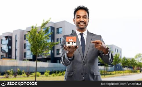 real estate business and people concept - indian man realtor with house model over living houses on city street background. indian man realtor with house model on city street