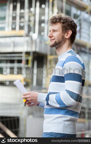 Real estate concept - young man on front of new big modern house building construction site with keys and blueprint project