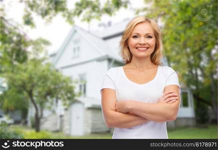 real estate, home and people concept - smiling woman in blank white t-shirt with crossed arms over private house background