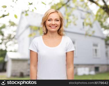 real estate, home and people concept - smiling woman in blank white t-shirt over private house background