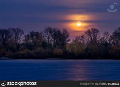 Real full moon over the autumn forest close to Dnieper river in Kiev, Ukraine. Soft clouds in the dark sky cover partially our natural satellite. The water looks like a glossy blue mirror.