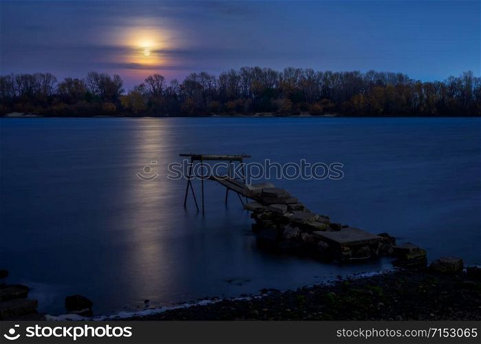 Real full moon over the autumn forest close to Dnieper river in Kiev, Ukraine. Soft clouds in the dark sky cover partially our natural satellite. A pontoon in the foreground. The water looks like a glossy blue mirror.