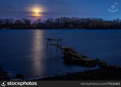Real full moon over the autumn forest close to Dnieper river in Kiev, Ukraine. Soft clouds in the dark sky cover partially our natural satellite. A pontoon in the foreground. The water looks like a glossy blue mirror.