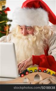 Real Santa Claus working on laptop with surprised face at home, surrounded by toys