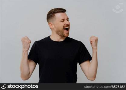 Real winner. Overjoyed young man in black t shirt raising clenched fists and screaming with excitment while reaching perconal goals, celebrating success triumph while standing against grey background. Overjoyed emotional happy man raising clenched fists and screaming while celebrating success