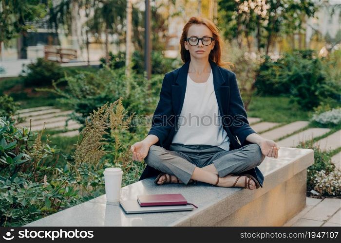 Realxed redhead woman does yoga outside at park sits in lotus pose meditates outdoors wears spectacles formal outfit gathers with thoughts before working breathes fresh air. Healthy lifestyle. Realxed redhead woman does yoga outside at park sits in lotus pose meditates outdoors