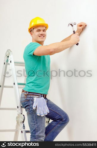 reapir, building and home renovation concept - smiling man in yellow protective helmet hammering nail in wall