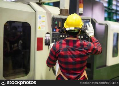 Rear Factory worker with plaid shirt and safety helmet command machine screen and keyboard to produce steel spare part by automatic drilling machine. Heavy industry with high technology.