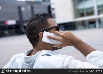 Rear image of a young man sitting on a city bench talking on the phone, wearing sunglasses.