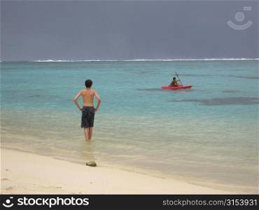 Rear of a young man standing on a beach watching a boat on the sea, Moorea, Tahiti, French Polynesia, South Pacific