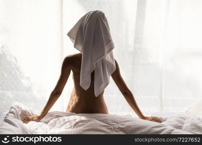 Rear of lonley woman wrapped shower towel after taking a bath sit naked on bed at window. Sexy nude tan girl stay at home due to covid-19 or coronavirus pandemic. Quarantine, social distancing.