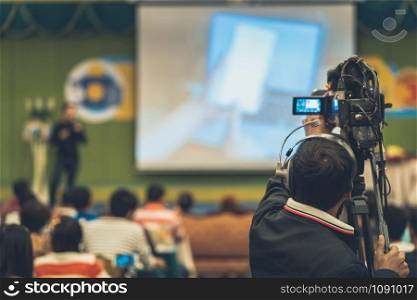 Rear side of Video Cameraman taking photograph to Asian Speaker with casual suit on the stage present the screen in the conference hall or seminar meeting, event and seminar production concept