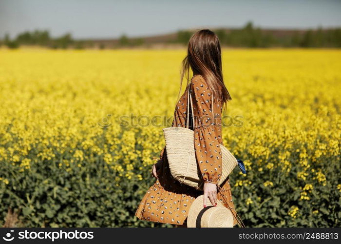 Rear view. beautiful young woman in a dress holding a hat and walking in a rapeseed field for summer, view from the back. copy space. summer holiday concept.. Rear view. beautiful young woman in a dress holding a hat and walking in a rapeseed field for summer, view from the back. copy space. summer holiday concept