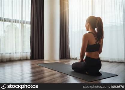 Rear view image of a beautiful young asian woman sitting on training mat preparing to workout at home