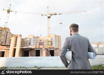 rear view male architect holding blueprint hard hat looking construction site