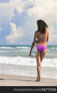 Rear view of a beautiful sexy young woman surfer girl in bikini with surfboard standing in the surf on a beach