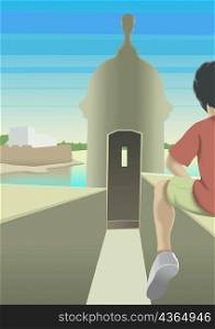 Rear view of a boy sitting on wall in front of a tower