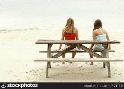Rear view of a girl and a teenage girl sitting on a picnic table on the beach