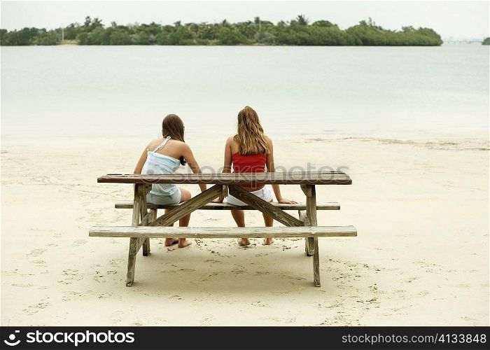 Rear view of a girl and a teenage girl sitting on a picnic table on the beach