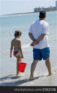 Rear view of a girl holding a bucket and walking with her father on the beach