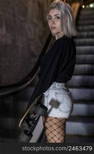 Rear view of a girl looking camera in shorts holding a long board while standing on mechanic stairs at night