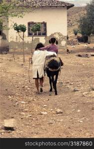 Rear view of a girl riding a donkey with her sister walking along her in a village, Turkey