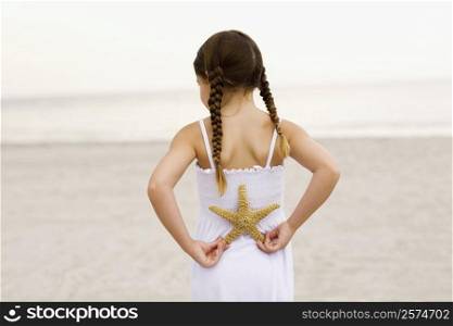 Rear view of a girl standing on the beach and holding a starfish behind her back