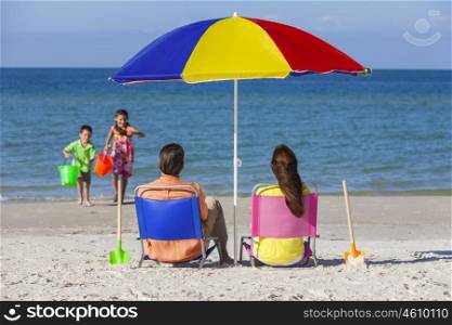 Rear view of a happy family of mother &amp; father, parents daughter &amp; son children having fun in deckchairs under an umbrella on a sunny beach