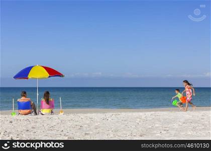 Rear view of a happy family of mother &amp; father, parents daughter &amp; son children having fun in deckchairs under an umbrella on a sunny beach