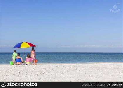 Rear view of a happy family of mother &amp; father, parents daughter &amp; son children sitting in deckchairs under an umbrella on a deserted sunny beach