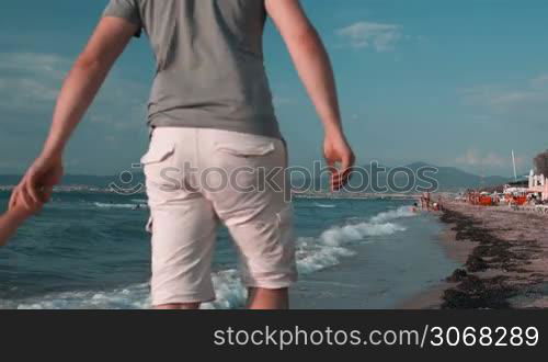 Rear view of a happy young Caucasian family, made of a man and his wife, both holding the hands of their child, while walking on the beach, in a beautiful summer day