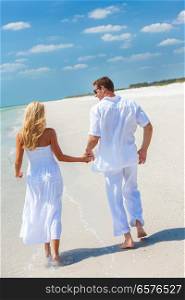 Rear view of a happy young man and woman couple running, laughing and holding hands on a deserted tropical beach with bright clear blue sky