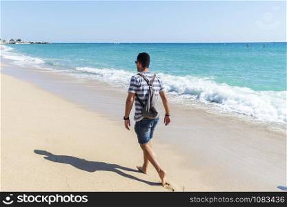 Rear view of a man with backpack walking away on seashore in a sunny day