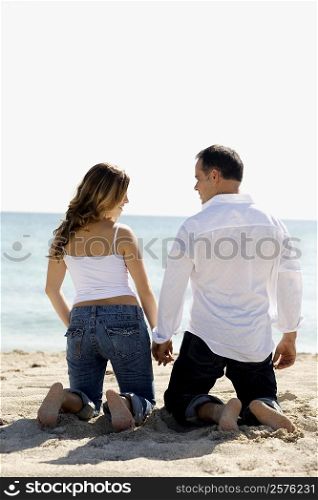Rear view of a mid adult man and a young woman kneeling with holding hands on the beach