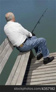 Rear view of a senior man sitting on a bench at the lakeside and fishing