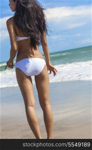 Rear view of a sexy young brunette woman or girl wearing a white bikini on a deserted tropical beach with a blue sky