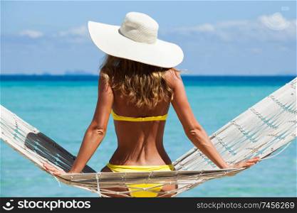 Rear view of a woman in bikini with a stylish hat sitting on a hammock on a beach by the sea. Woman in hammock on beach