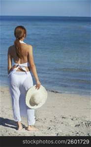 Rear view of a woman standing on the beach and holding a straw hat