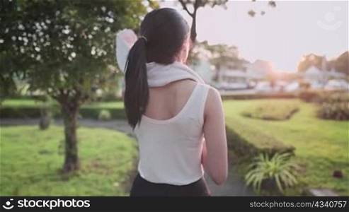 Rear view of a young athletic healthy skin woman walks on run way to cold down after work out and wipes out sweat using white towel on her neck in green beautiful park, healthy and lifestyle