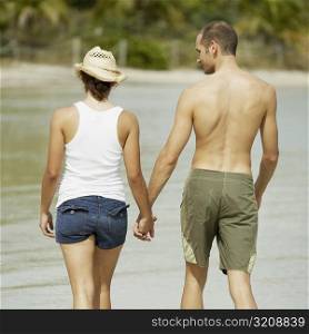 Rear view of a young man and a teenage girl holding hands and walking on the beach