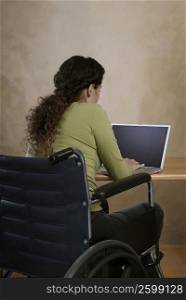 Rear view of a young woman sitting in a wheelchair and using a laptop