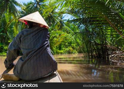 Rear view of anonymous local woman wearing traditional leaf hat paddling a canoe on the Mekong Delta, Vietnam, South East Asia
