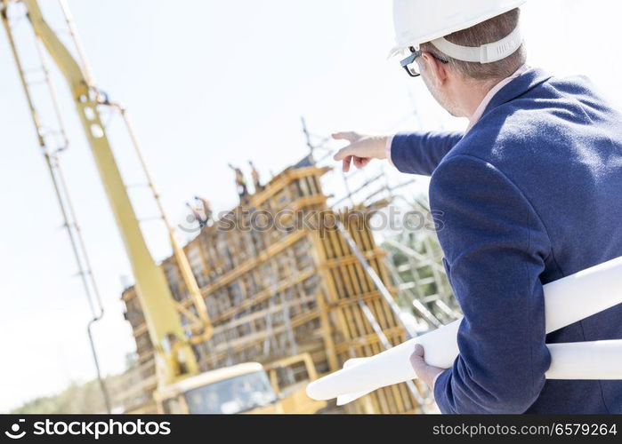 Rear view of architect holding blueprints while pointing at construction site