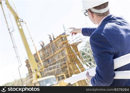 Rear view of architect holding blueprints while pointing at construction site
