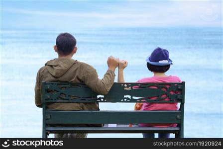 Rear view of Asian adult couple on metal bench showing pinky swear hand sign to be a relationship forever after dating on the beach, travel and love concept