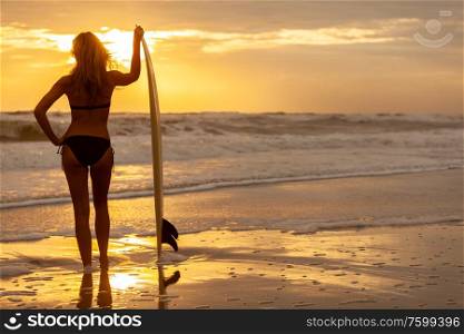 Rear view of beautiful sexy young blonde woman surfer girl in bikini with white surfboard on a beach at sunset or sunrise
