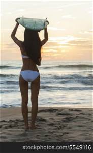 Rear view of beautiful sexy young woman surfer girl in bikini with white surfboard on her head on a beach at sunset or sunrise