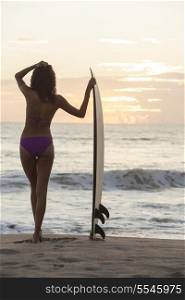 Rear view of beautiful sexy young woman surfer girl in purple bikini with white surfboard on a beach at sunset or sunrise
